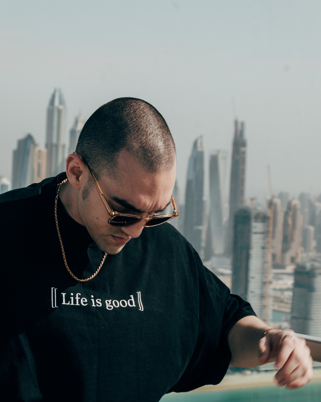 Male wearing a black tshirt that read life is good with a gold chain around his neck and dubai marina in the background